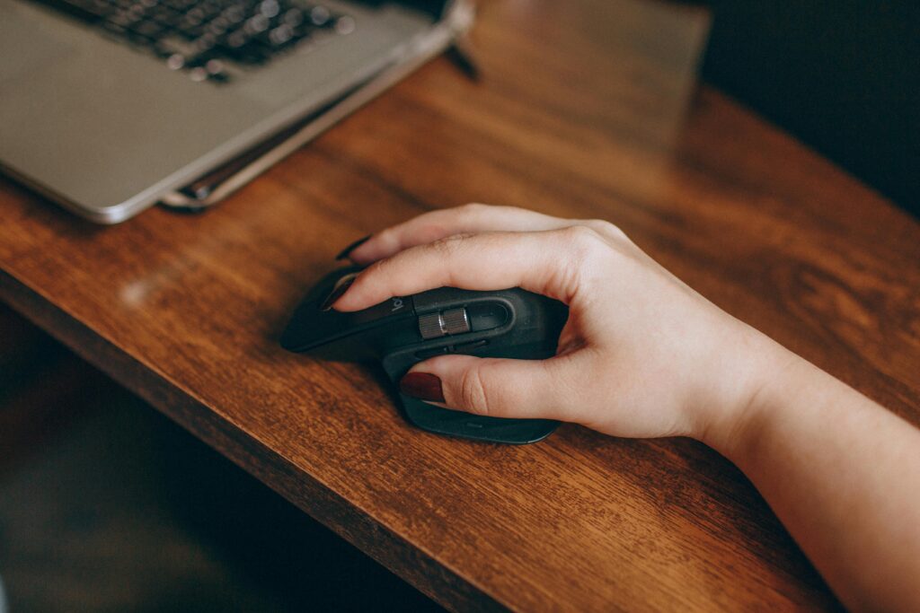 Dłoń klikająca myszkę przy laptopie na drewnianym biurku – clickbait co to znaczy, czyli chwytliwy nagłówek zachęcający do kliknięcia poprzez wzbudzanie ciekawości lub silnych emocji.